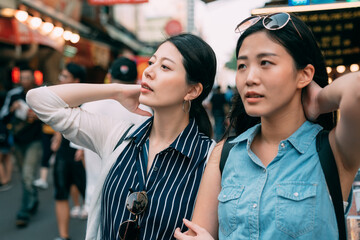 closeup two attractive japanese women are touching their hair and looking at the distance while shopping together on a hot sweaty summer day in downtown area.