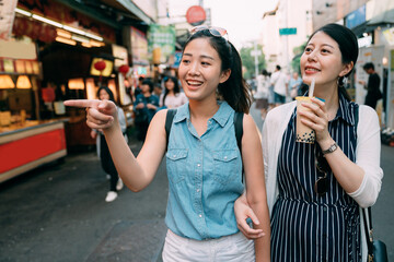 happy asian girl is pointing at distance and suggesting her friend with bubble milk tea to check out while shopping together on a sightseeing street in summer