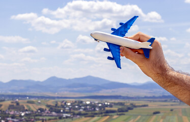 man hand holding airplane against the sky,mountains landscape background.sunny summer day,vacation,tourism,travel,trip concept.blue and white color plastic toy plane.fly,book ticket,adventure