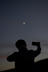 vertical portrait of a young man taking a picture of the Moon with his cell phone at sunset at dusk.