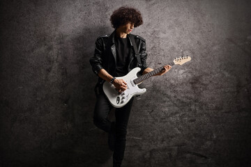 Male musician in a leather jacket with an electric guitar leaning on a dark rugged wall