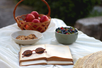 Picninc blanket with straw bag, bowl of strawberries and blueberries, bowl of chocolate chip cookies, books, sunglasses and basket of apples in the garden. Selective focus.