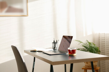Home office concept. Designated work from home area near the window. Modern laptop with blurred screen and glass cup of tea on table. Close up, copy space, interior background.