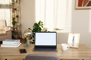 Blank screen laptop, cell phone and a cup on wooden table. Cozy lofty office with house plants full of natural sunlight. Close up, copy space, background.