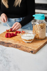 Yogurt with granola, and raspberries and strawberries next to jar with granola in a bright modern kitchen and a woman cutting strawberries