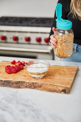 Yogurt with granola, and raspberries and strawberries and a female holding jar with granola in a bright modern kitchen