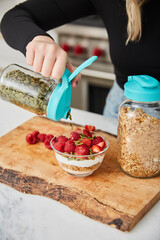 A woman topping yogurt, granola, fruit bowl with pumpkin seeds in a bright modern kitchen
