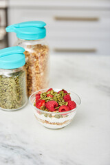 Yogurt with granola topped with raspberries and strawberries next to jars with granola and nuts in a bright modern kitchen
