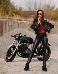 Naklejka na ściany i meble beautiful girl with long dark hair red lips in black jacket and black pants standing near black vintage motorcycle in industrial zone