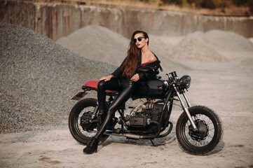 Plakat beautiful girl with long dark hair red lips in a black jacket and black pants sits on a black vintage motorcycle in an industrial zone