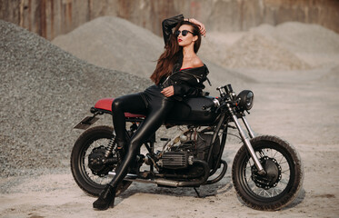 Obraz na płótnie Canvas beautiful girl with long dark hair red lips in a black jacket and black pants sits on a black vintage motorcycle in an industrial zone