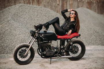 
beautiful girl with long dark hair red lips in a black jacket and black pants sits on a black vintage motorcycle in an industrial zone