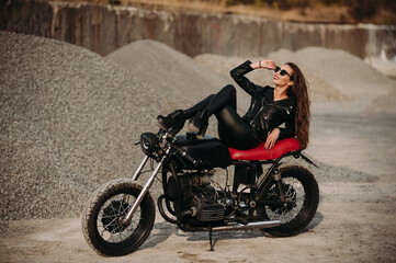 Naklejka na ściany i meble beautiful girl with long dark hair red lips in a black jacket and black pants sits on a black vintage motorcycle in an industrial zone
