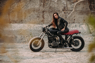 Fototapeta na wymiar beautiful girl with long dark hair red lips in a black jacket and black pants sits on a black vintage motorcycle in an industrial zone