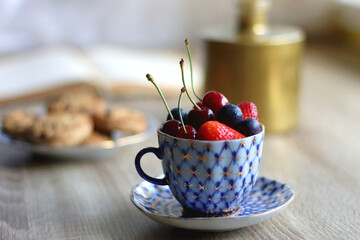 Plate of chocolate chip cookies, cup filled with strawberries, blueberries and cherries, open book and vase with gypsophila flowers on the table. Selective focus.
