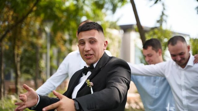Groom flexing and having fun walking with groomsmen on wedding day. Happy classy man in black suit, bow tie and boutonniere hanging out partying with friends and cheering posing at camera in a park.
