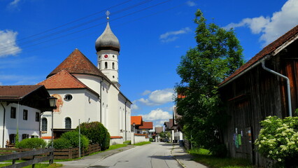 Fototapeta na wymiar idyllische Dorfstraße in Unterammergau in Bayern mit Kirche, Stall, Baum und Häusern unter blauem Himmel