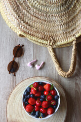 Round straw bag, brown sunglasses, pink earphones and bowl of berries on wooden table. Flat lay.