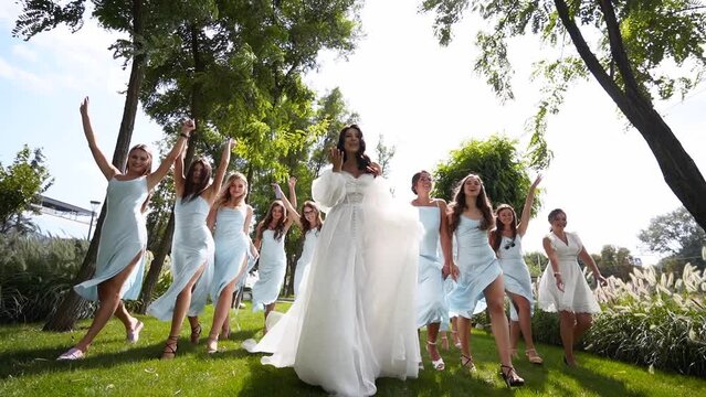 Beautiful bride and her pretty bridemaids in pale blue dresses walking in a park cheering waving hands. Woman with long airy bridal veil in elegant white dress party, hug with friends. Slow motion.