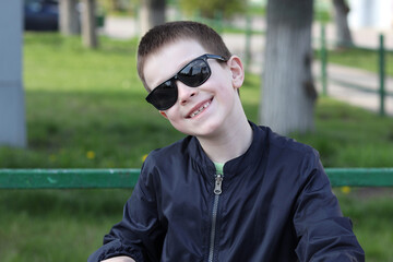A 7-year-old boy in sunglasses, smiling happily on a sunny bright day against a background of green trees