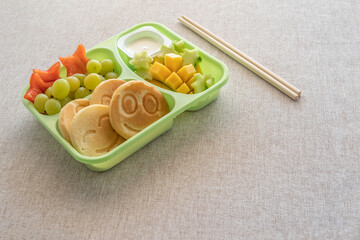Lunch box with pancakes, mango fruit, cucumber, grapes for kid's breakfast on cotton fabric background.