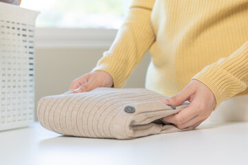 Fototapeta premium Housewife, asian young woman hand in many folding freshly shirts, sweaters or dress on desk, table after washing clean clothes and drying, making household working in room at home. Laundry and maid.