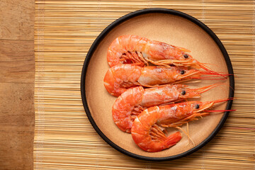 Four cooked red tiger shrimps on a beige rustic plate on bamboo matt from above.