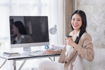 Portrait of Attractive Asian woman working with laptop computer in the office hand holding cup of coffee.