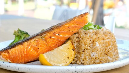 Close up of fried salmon steak with vegetables, lemon and rice on white plate. Healthy lunch served in restaurant. Portion of cooked fish fillet. Delicious seafood with resort on background