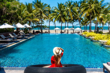 Luxury beach vacation in tropical beach hotel. Tourist woman in red dress relax near blue swimming pool in modern resort. Female traveler on sea vacation. - obrazy, fototapety, plakaty