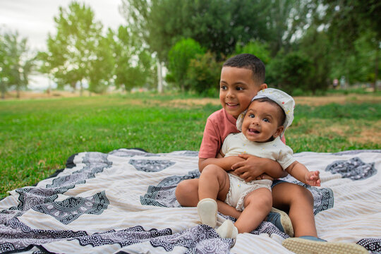 Brother holding his baby sister in his arms, sitting on the grass in the park, image with copy space
