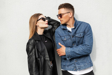 Fashionable beautiful hipster couple with cool vintage sunglasses wearing trendy casual denim outfit with blue jeans jacket and black leather coat standing near the wall on the street. Man and woman