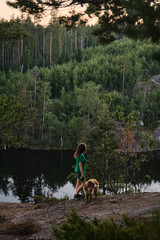 Young woman stands on rock with dog and admires nature and river below. The concept of traveling in Russia. Australian Shepherd travels to Karelia in summer with owner.
