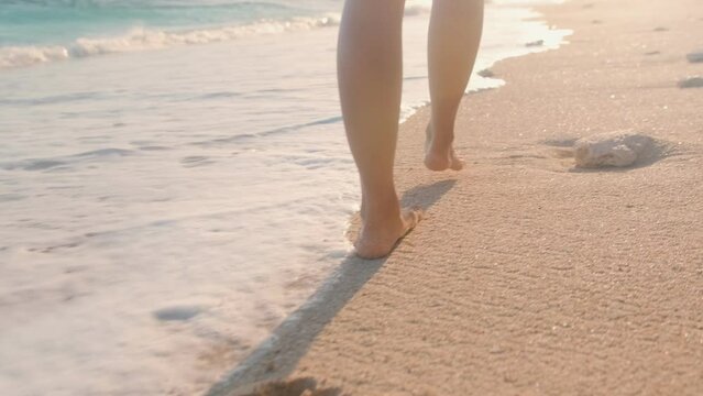 Close up beautiful bare feet woman walking on sea sand at high tide passing next to waves of ocean leaving footprints on ground be on sunny beach. Summer holidays, recreation, tourism