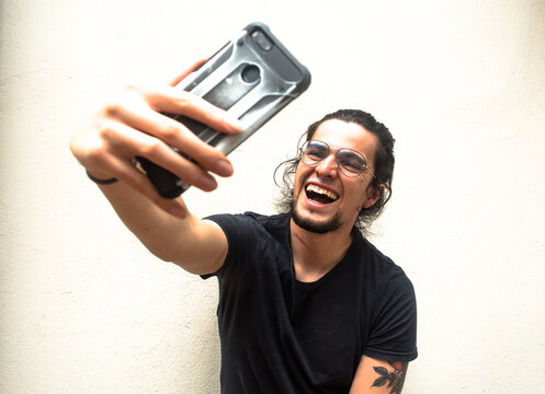 Portrait of long haired tattooed man with beard holding a phone on his hand smiling and laughing taking a selfie. White background