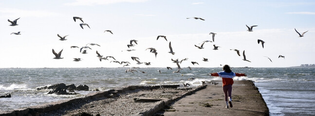 Chica corriendo, asustando y espantando a los pájaros gaviotas en una rampa en el mar con rojas y oleaje