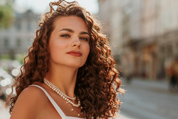 Beautiful confident curly brunette woman wearing trendy pearl layered necklace, posing in street of...