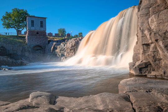 Waterfall over quartzite red rocks at Big Sioux River Park in Sioux Falls, South Dakota. Long exposure photo of the silky flowing water with the view of the Queen Bee Turbine House on the hilltop.