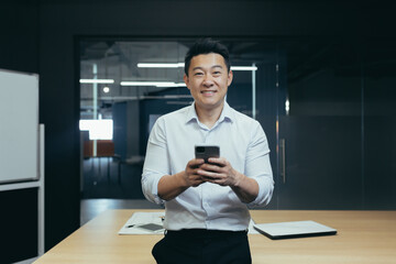 Portrait of a successful Asian businessman, man at work, standing and typing on the phone, smiling...