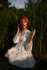 Sensual pretty woman with short red hair sits on the ground among grass, poses and looks at the sunset. Portrait of a cute red haired girl in a white dress as poster, wallpaper or advertising design.