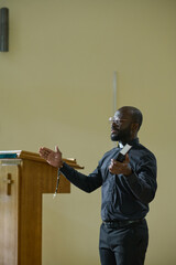 Young confident pastor in black shirt with clerical collar and trousers blessing pashioners while...