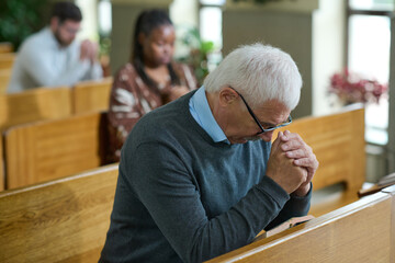 Mature man inclining his head and bending over his hands put together during silent pray after sermon or before communion in church
