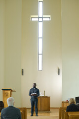 African American priest in black casualwear making sermon for parishioners while standing against...