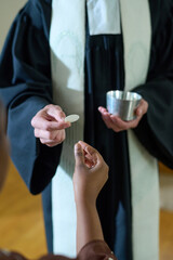 Hand of young Catholic priest in cassock passing unleavened bread to African American woman during communion rite in church