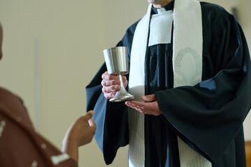 Clergyman of Catholic church holding cup with wine for oblation rite while standing in front of...