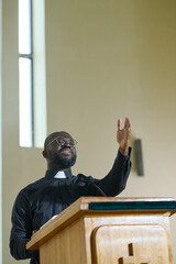Young priest in black shirt with clerical collar standing by pulpit and speaking in microphone...