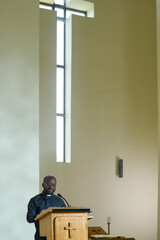 Young black man standing by wooden pulpit in front of parishioners while reading Bible aloud and...