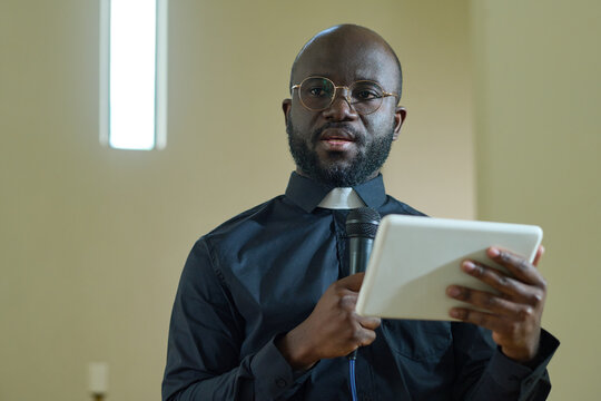 Young confident black man with microphone and digital tablet making speech for parishioners of evengelical church during sermon