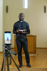 African American pastor in black pants and shirt with clerical collar holding open Bible while standing in front of smartphone camera - obrazy, fototapety, plakaty