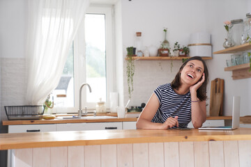 A happy woman is in the kitchen in her house. The woman laughs happily and looks away. There is a laptop next to her. The concept of remote work. High quality photo